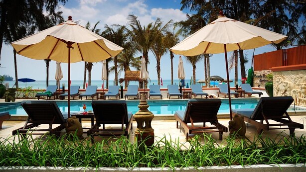 A poolside area with lounge chairs and umbrellas, palm trees, and a view of the ocean. - Kamala Beachfront Apartment (Kamala)