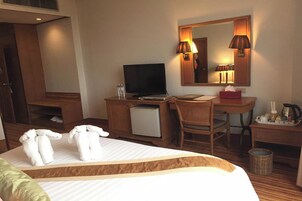 Habitación de hotel con cama, escritorio con una laptop, televisor, silla, refrigerador pequeño, espejo y mesita de noche con lámpara y caja de pañuelos.