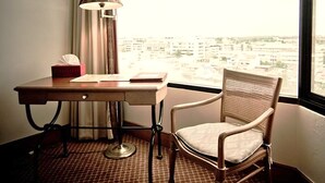 A hotel room with a desk, chair, lamp, and a view of the city through the window.