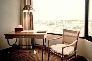 A hotel room with a desk, chair, lamp, and a view of the city through the window.