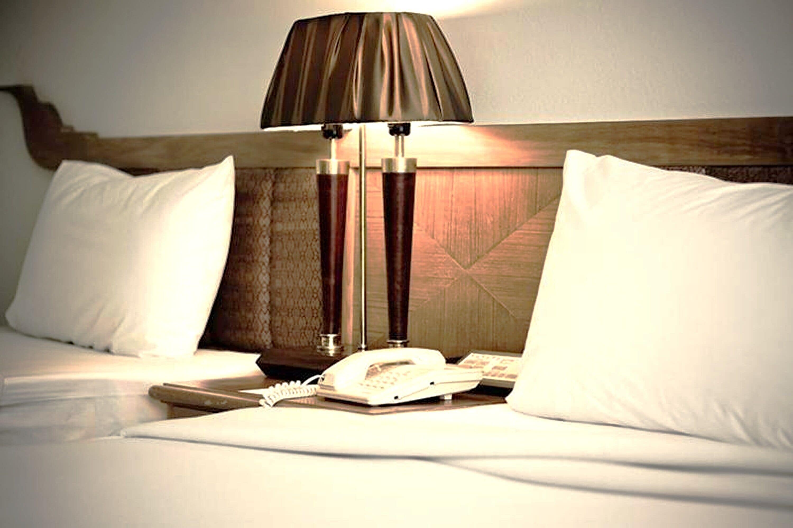 Habitación de hotel con cama, almohadas blancas, lámpara de noche, teléfono y cabecero de madera.