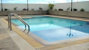 A rectangular swimming pool with a metal ladder on one side, surrounded by a tiled deck and a low wall with plants.