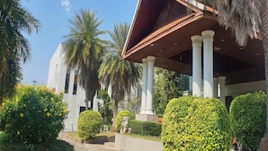 Um edifício com entrada coberta, colunas brancas e vegetação exuberante, incluindo palmeiras e arbustos.