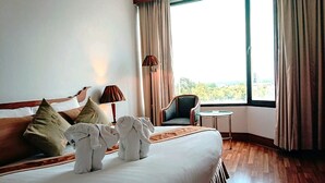 Quarto de hotel com uma cama grande, duas almofadas, mesa de cabeceira com abajur, uma cadeira e vista para o exterior pela janela.