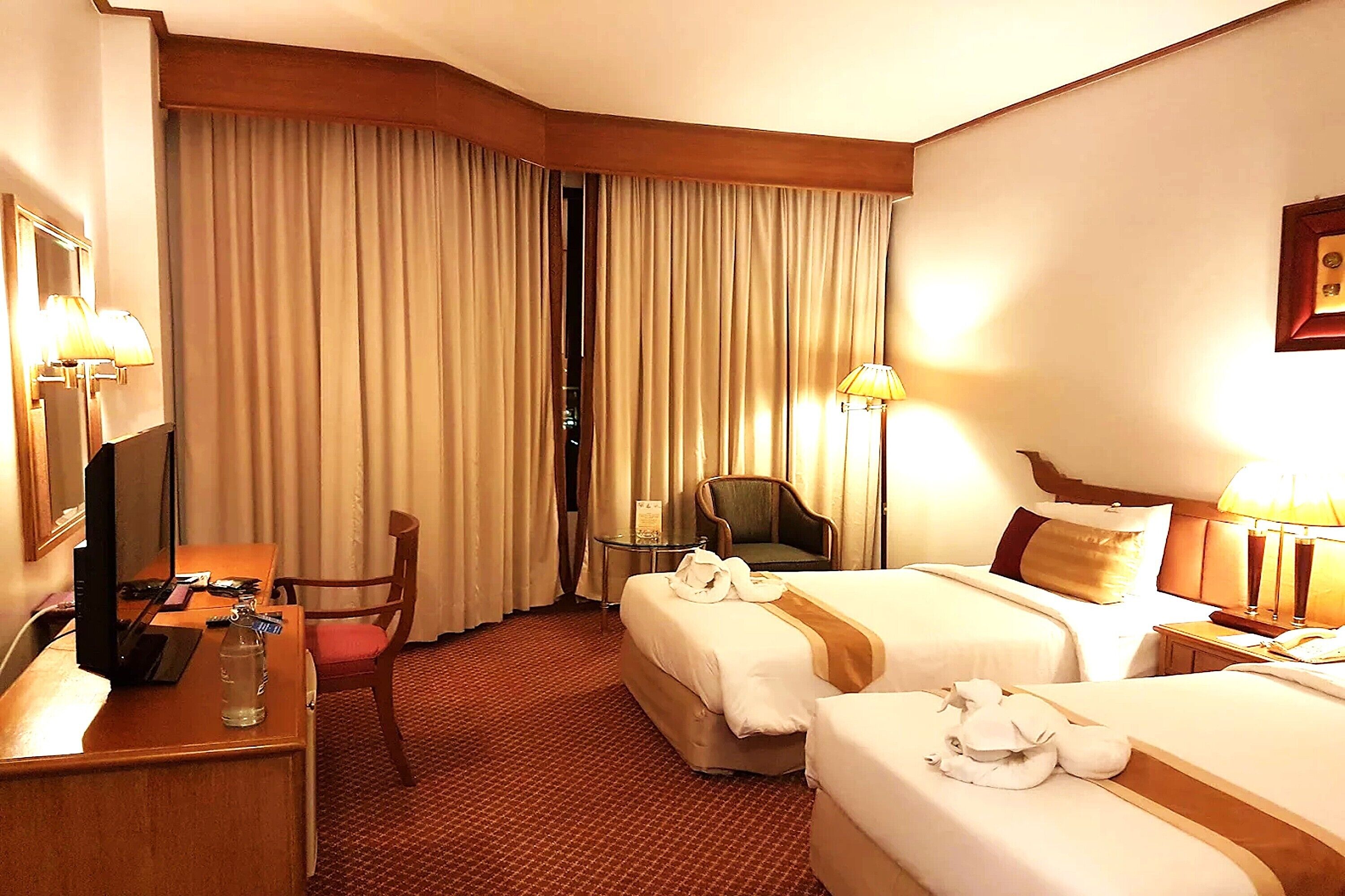 Une chambre d’hôtel avec deux lits, un bureau avec un ordinateur, une chaise, une petite table avec une lampe et une fenêtre avec des rideaux.