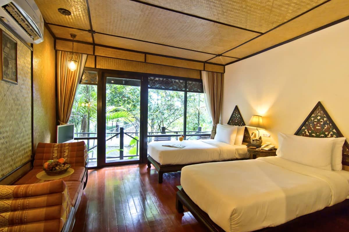 A hotel room with two beds, a sofa, a dining table, and a large window with a view of greenery.