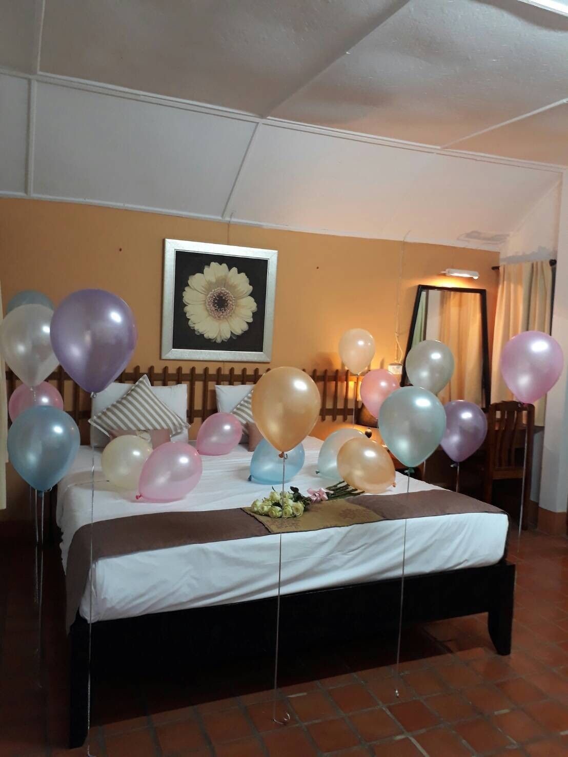 A hotel room with a bed, balloons, and a framed flower artwork on the wall.