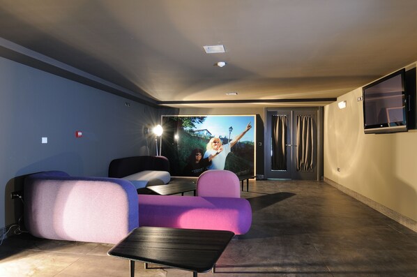 A modern hotel room with a large flat-screen TV mounted on the wall, a seating area with a pink sofa and a small table, and a door leading to another room.