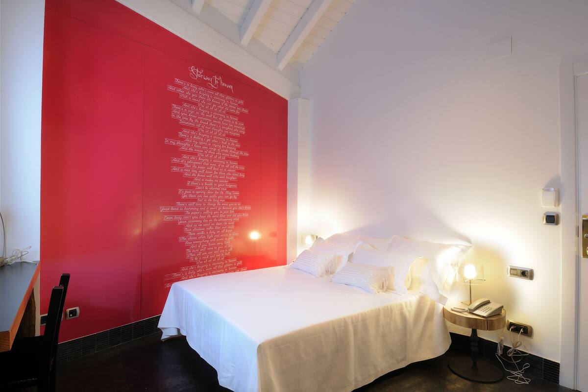 A hotel room with a large red wall, a bed with white linens, a bedside table with a lamp, and a wall-mounted phone.