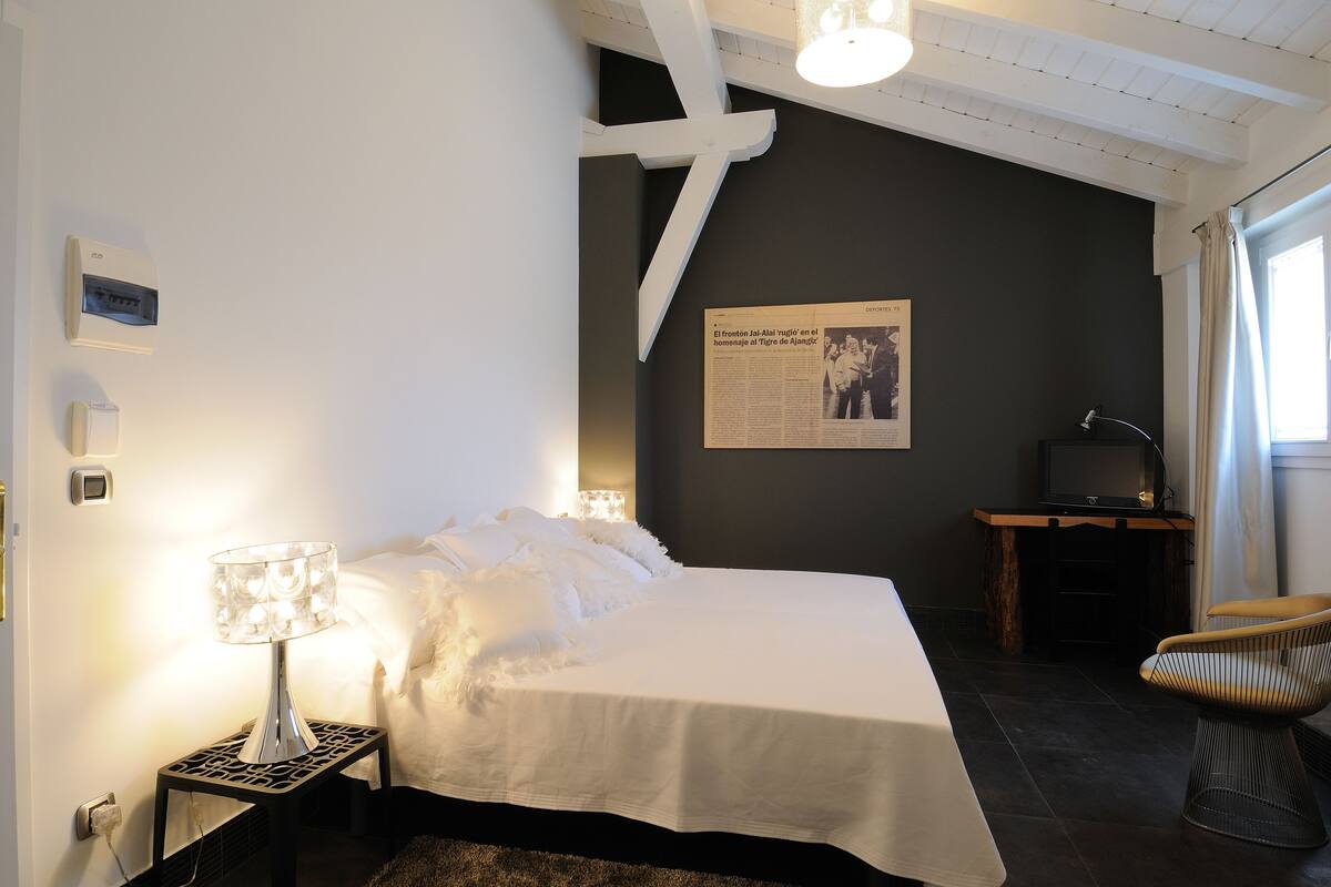 A bedroom with a bed, a nightstand with a lamp, a chair, a desk with a computer, and a poster on the wall.