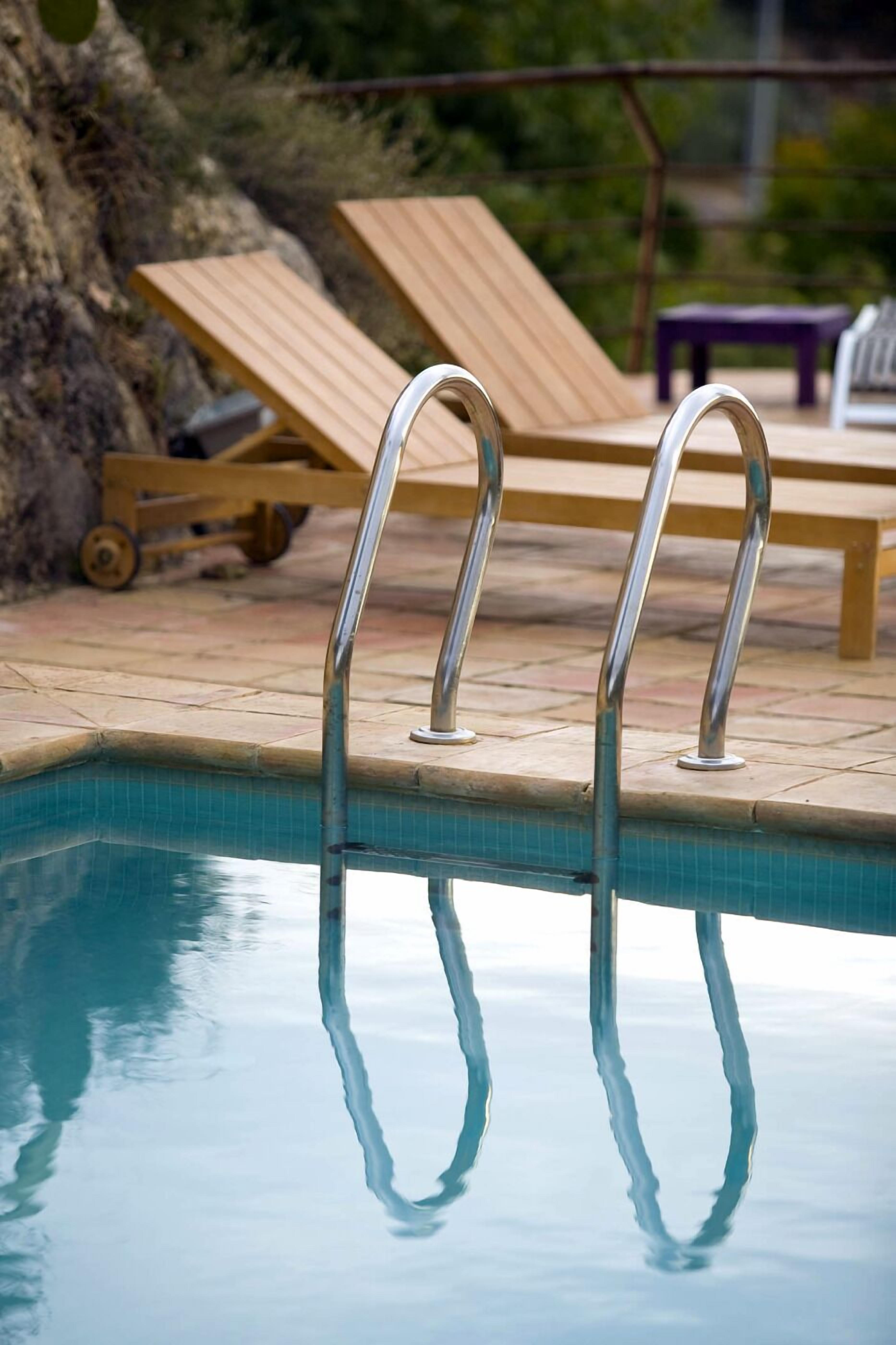 Una piscina con escaleras de metal, sillas de descanso de madera y un muro de piedra al fondo.