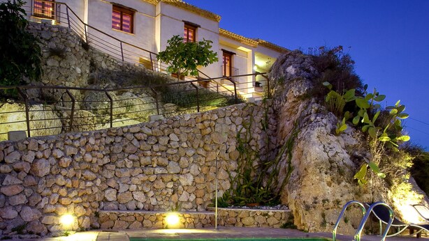 A swimming pool with a stone wall and a building with red windows in the background. -  Hospedería Bajo el Cejo  (Alhama de Murcia)