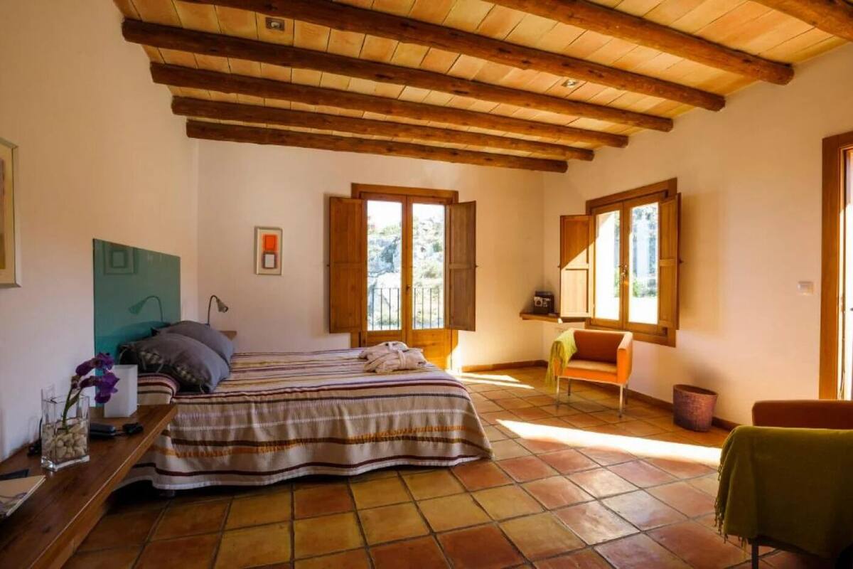 A bedroom with a bed, a chair, a table, and a window with shutters.