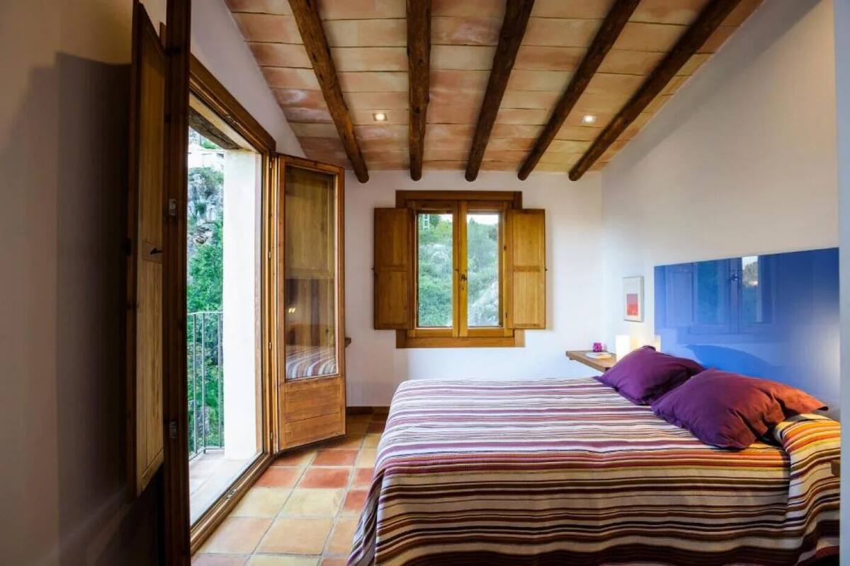A bedroom with a large bed, wooden beams on the ceiling, a window with wooden shutters, and a view of greenery outside.