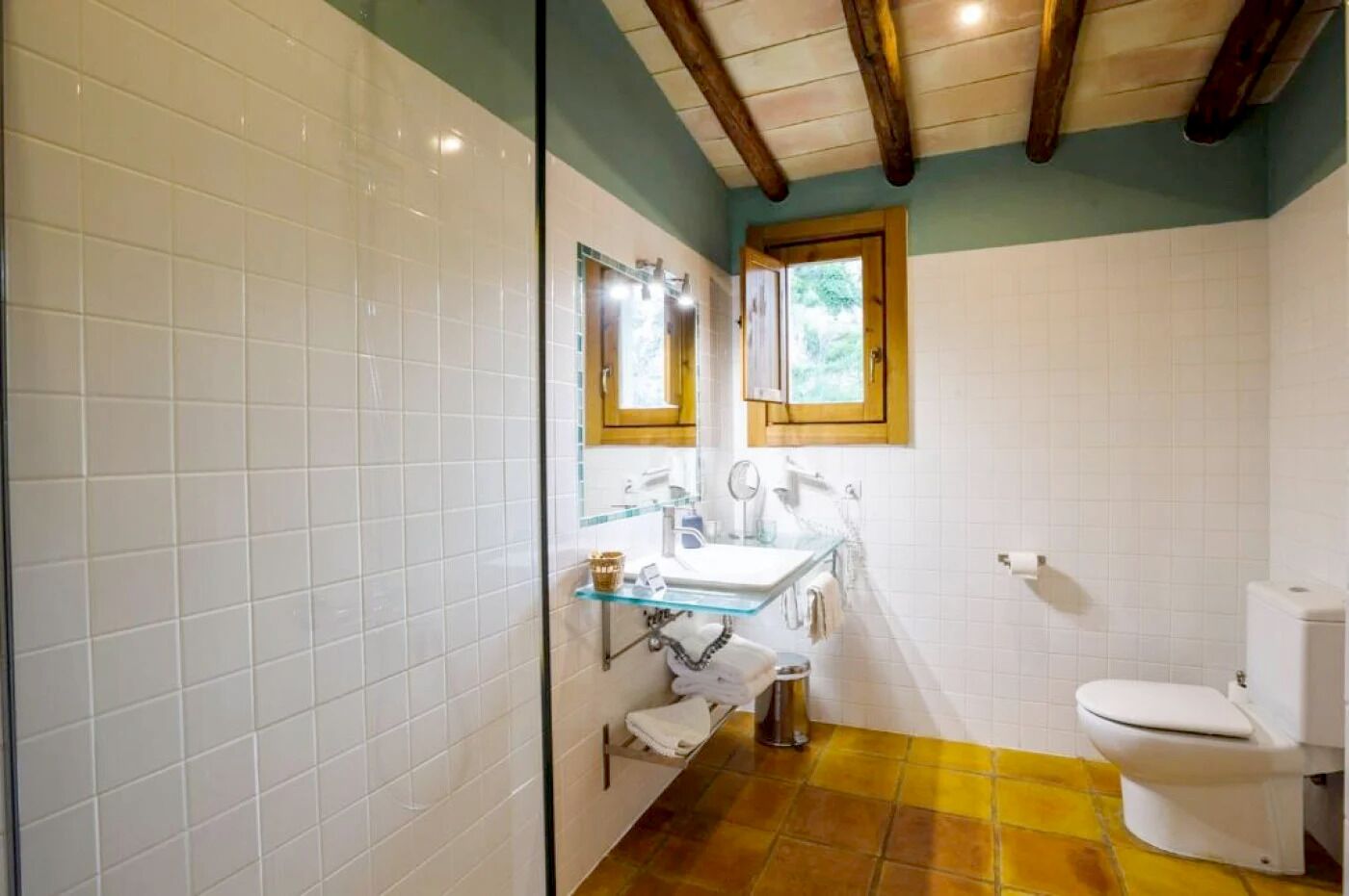 A bathroom with white tiled walls, a glass shower enclosure, a wooden ceiling, a white toilet, and a yellow-tiled floor.