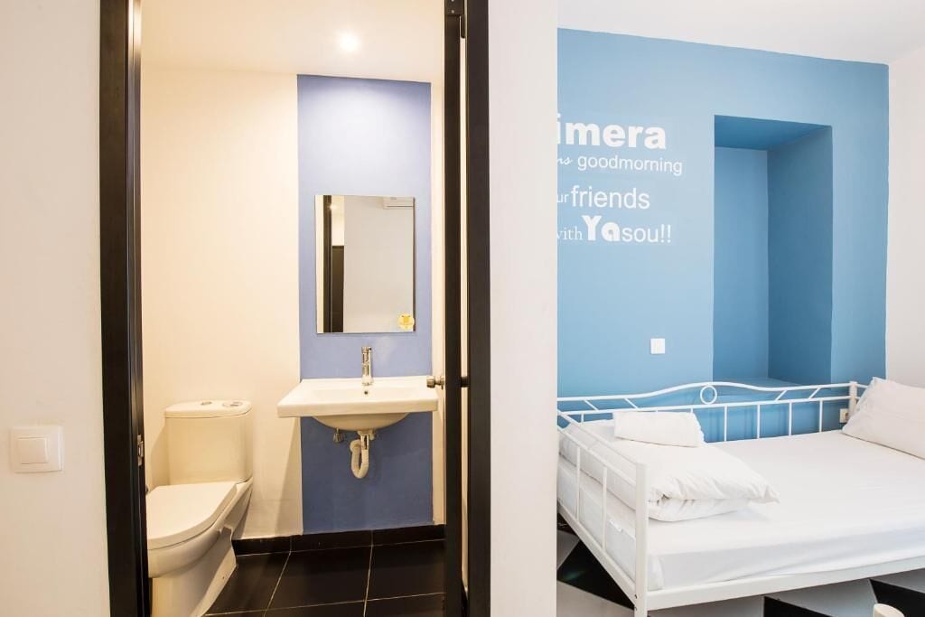 A modern bathroom with a toilet, sink, and mirror. The room has a blue accent wall and a bed with white bedding.