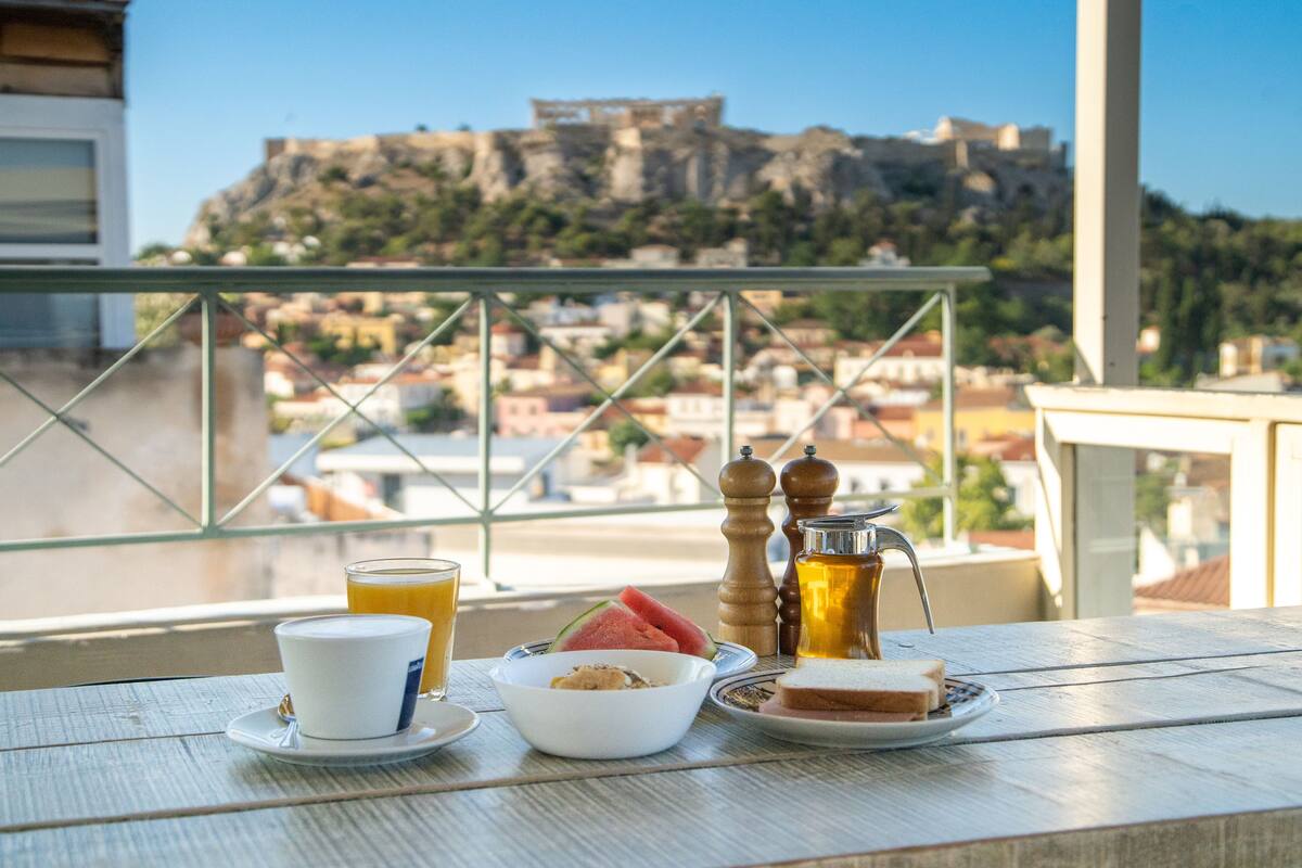 Mesa en balcón con taza, plato, tazón y jarrón de bebida, con vista a un edificio histórico.