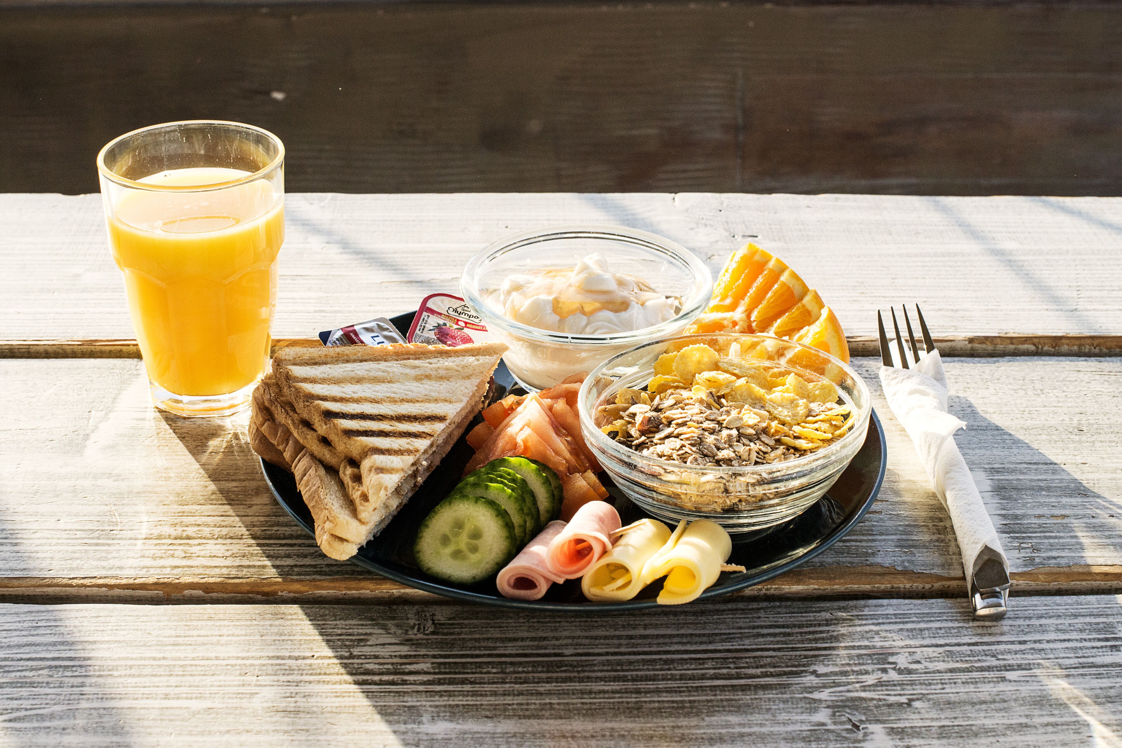 A breakfast plate with toast, ham, cheese, cucumber, and a bowl of cereal.