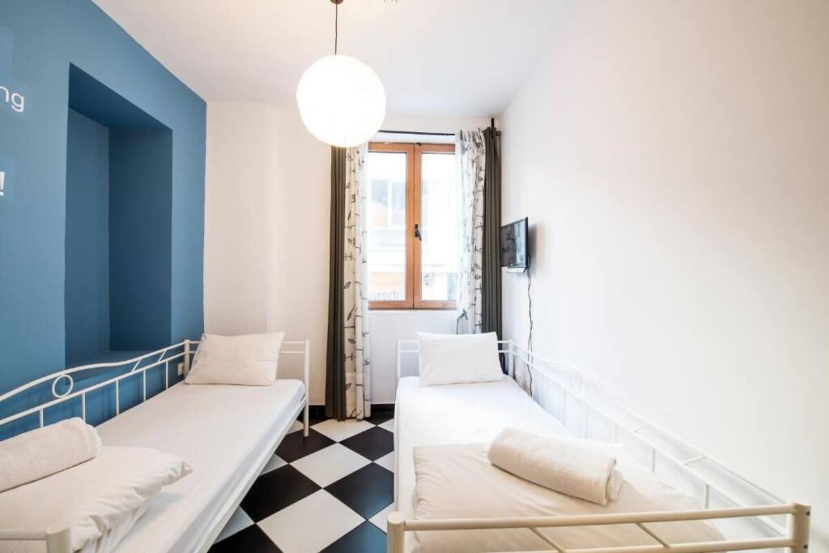 Una habitación con dos camas individuales, piso a cuadros, pared azul de acento, lámpara colgante y una ventana con cortinas.
