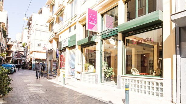 A street with shops, a blue car parked on the side, and a person walking. - Safestay Athens Monastiraki (Athens)