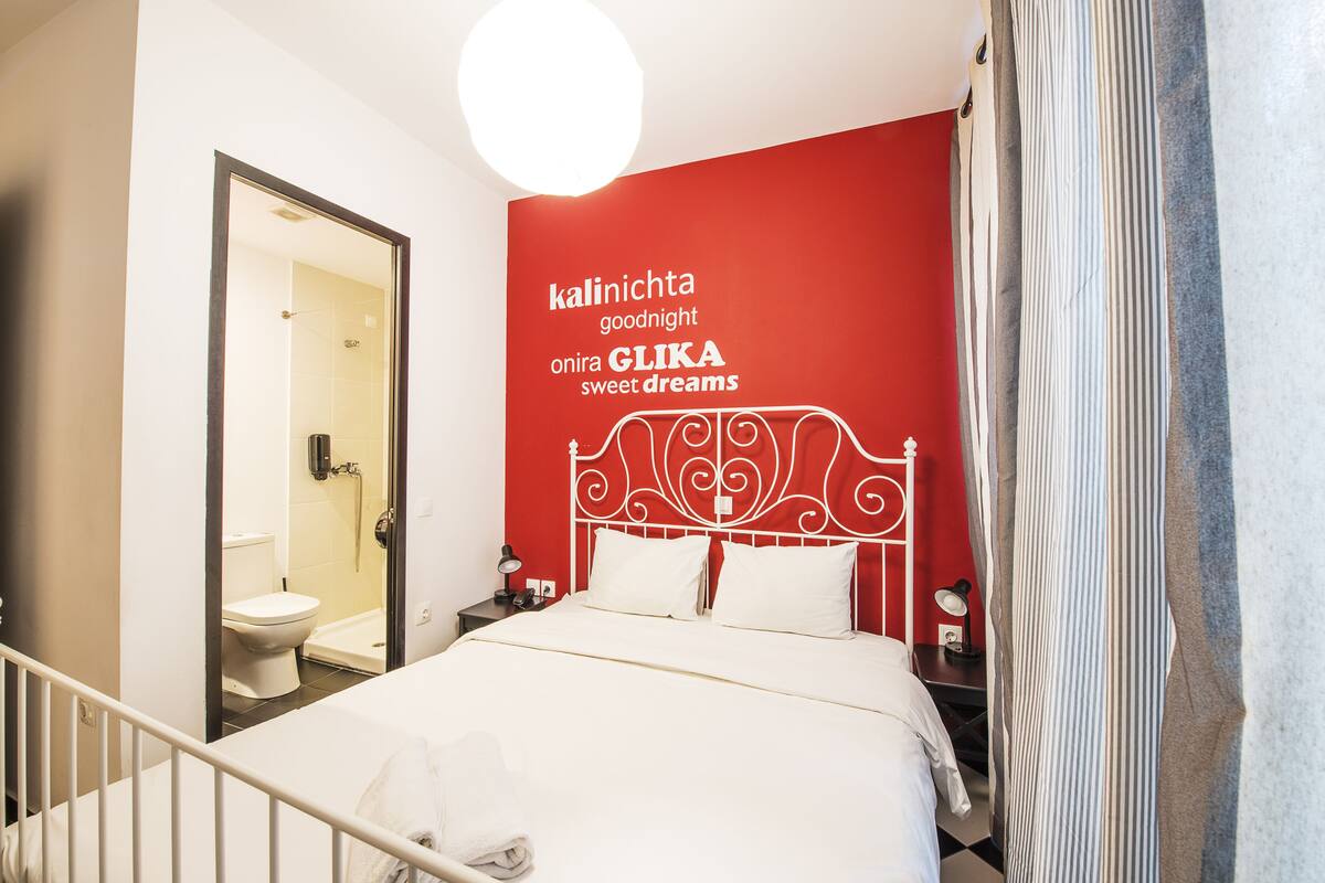 Habitación de hotel con una pared de acento roja, una cama blanca y un baño visible a través de una puerta abierta.