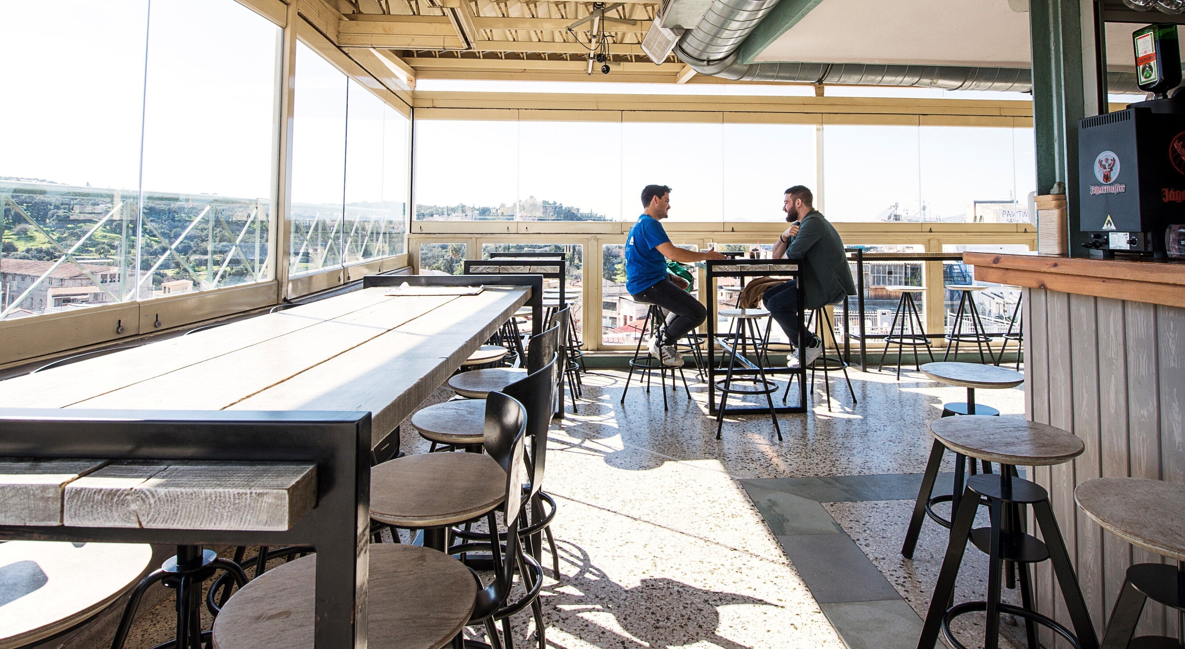 Un área de comedor al aire libre con mesas y sillas, ventanas amplias con vista a la ciudad y una barra con grifo de cerveza.