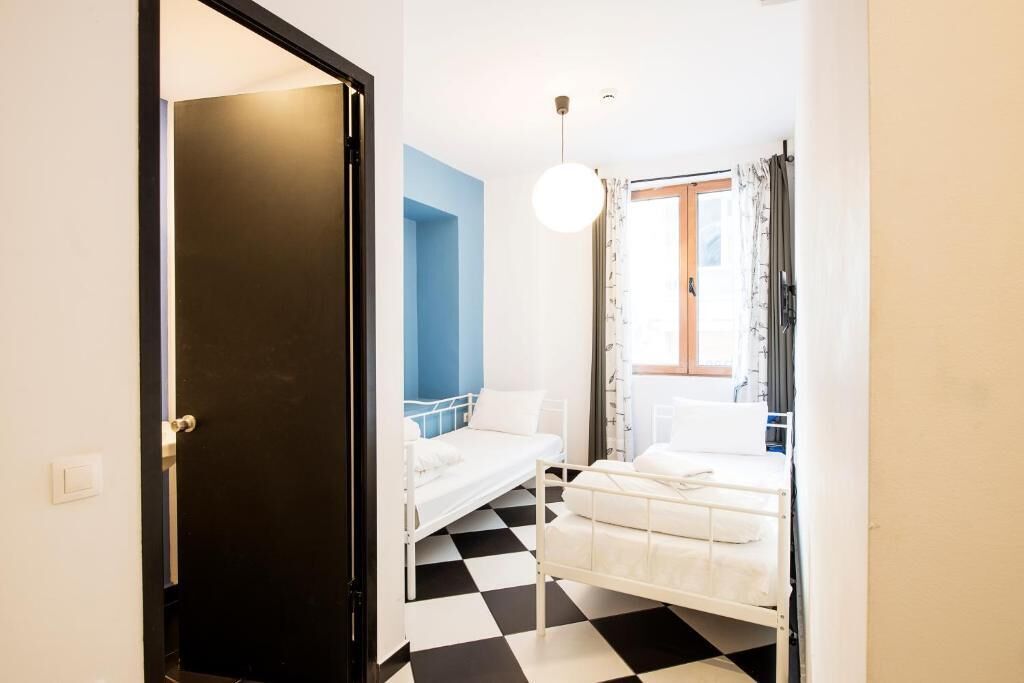Een kamer met twee bedden, een zwart-wit geruit vloerkleed, een blauwe accentmuur en een raam met gordijnen.