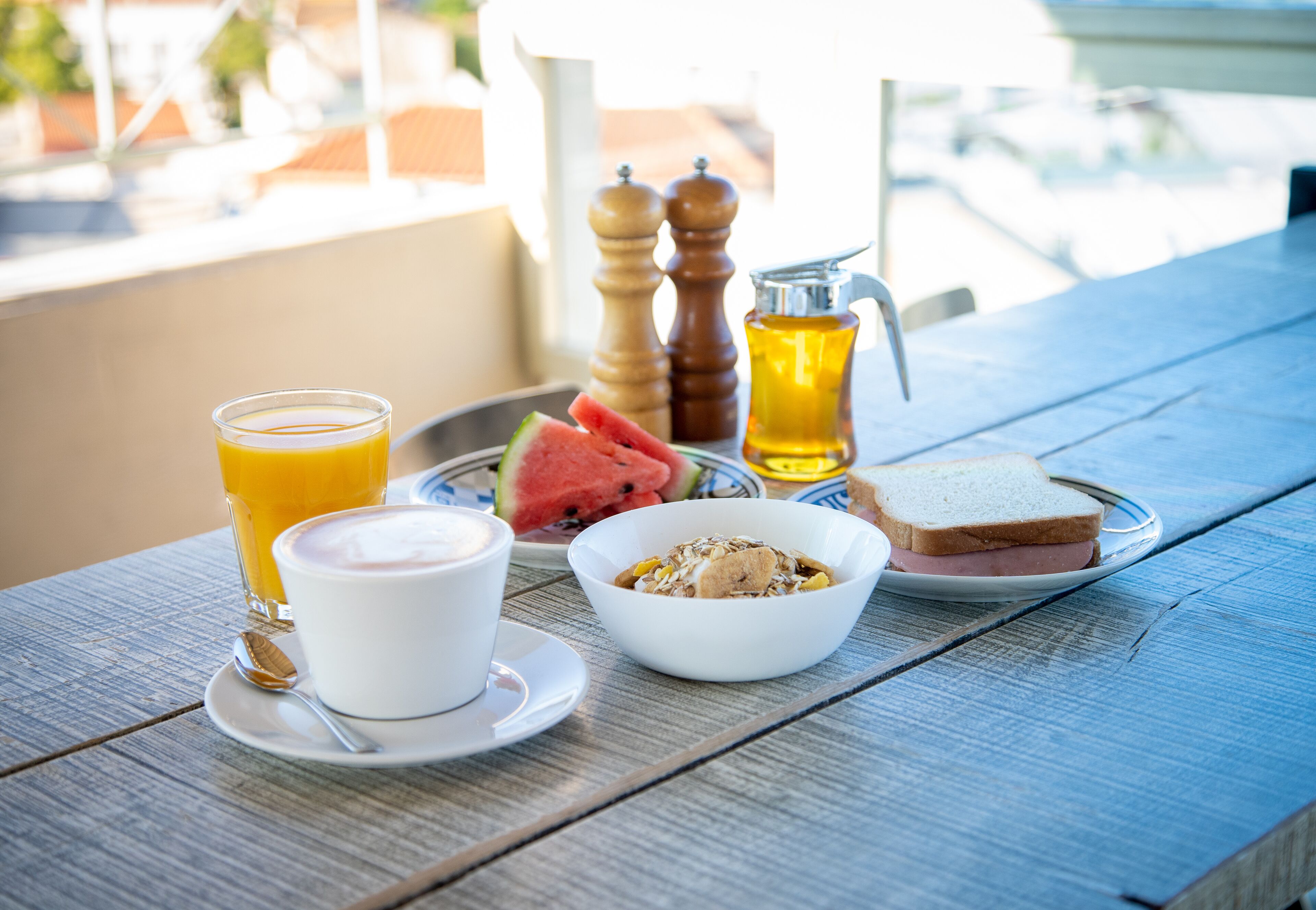 Bandeja de desayuno con vaso de jugo de naranja, taza de café, tazón de cereal, rebanada de pan y porción de sandía sobre una mesa de madera.
