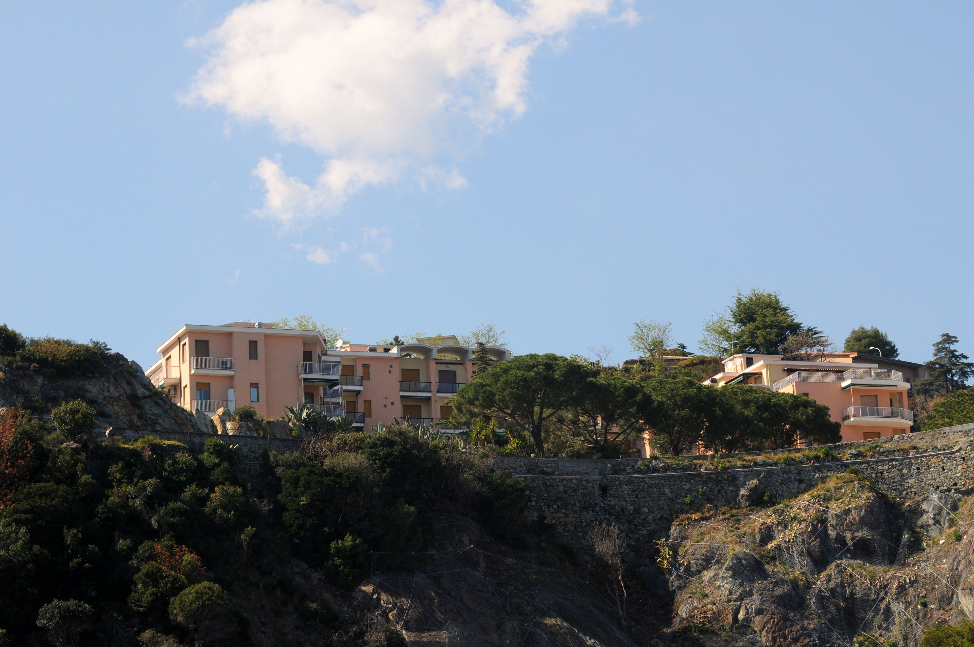 A coastal residential area with multi-story buildings, some with balconies, set against a backdrop of rocky terrain and sparse vegetation.