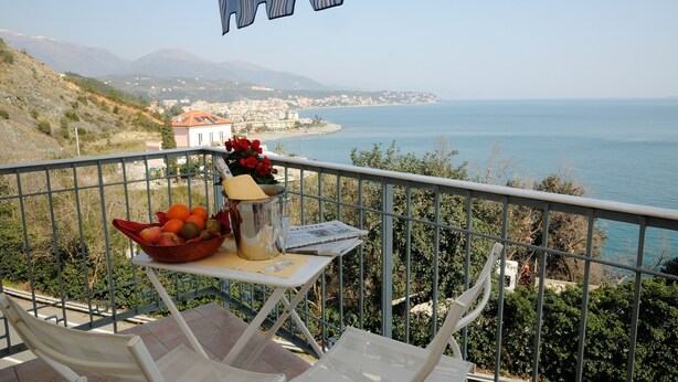 A balcony with a table holding a fruit bowl, a glass, and a notebook. The balcony overlooks a coastal view with mountains in the distance. - Hotel Puntabella (Varazze)