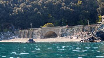 A stone bridge over water, leading to a rocky shore with a forested hill in the background.