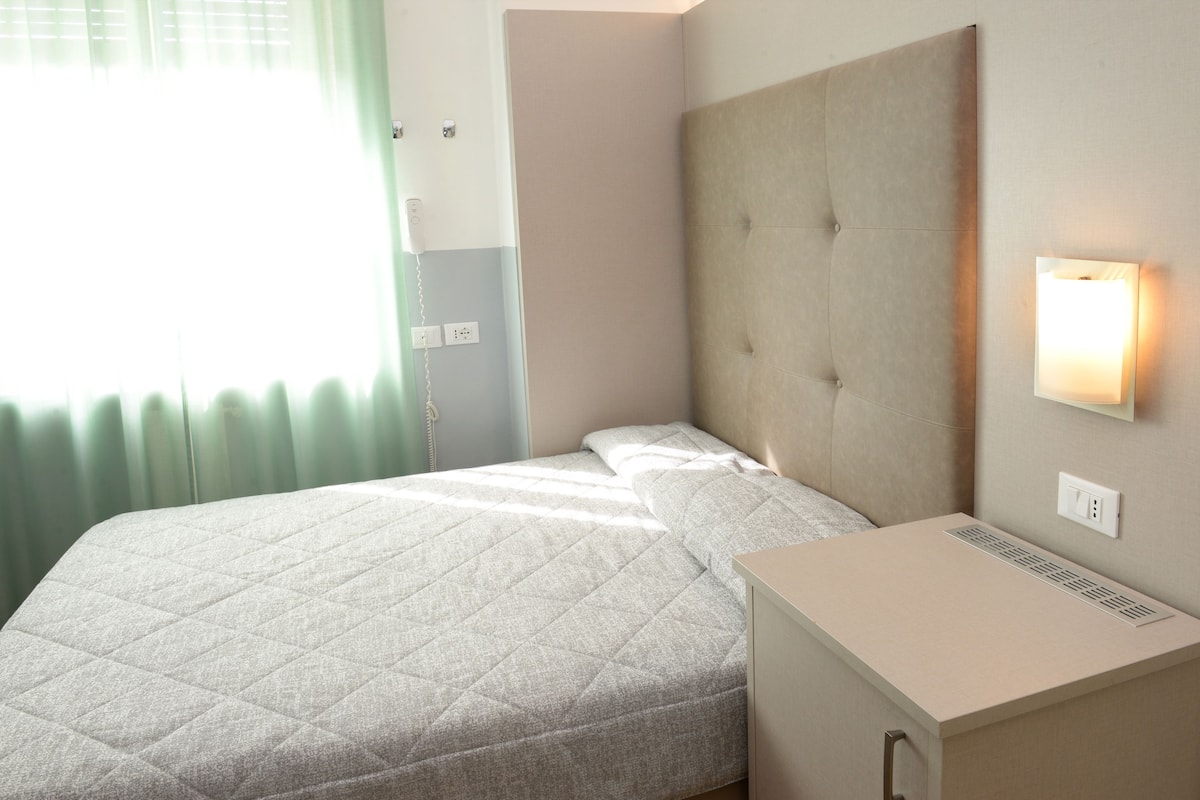 A neatly made bed with a quilted cover, a bedside table with a lamp, and a power outlet on the wall.