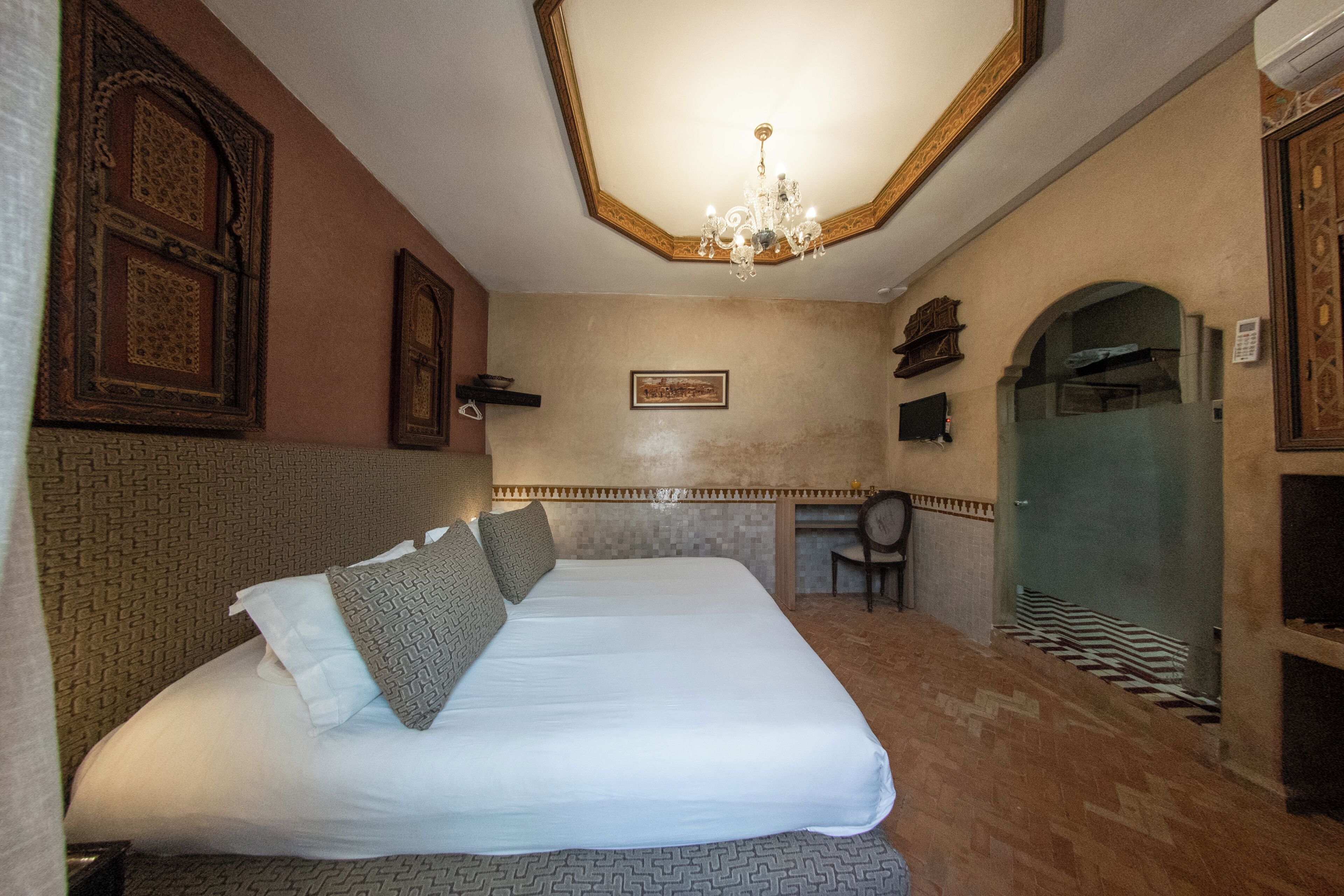 A hotel room with a large bed, a chandelier, a shower, and a small desk.