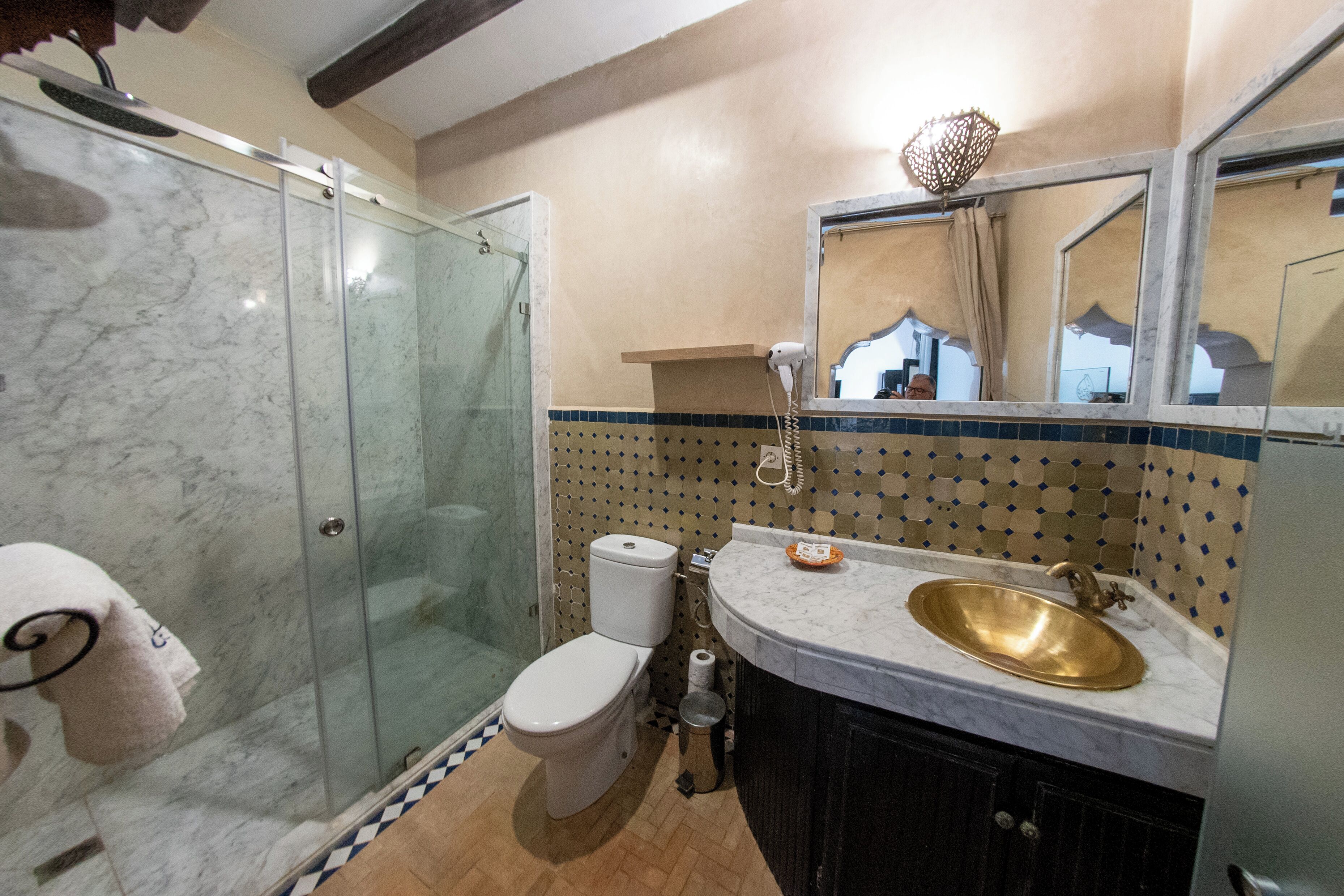 A bathroom with a glass shower enclosure, a toilet, a sink with a gold basin, and a mirror with a decorative light fixture.