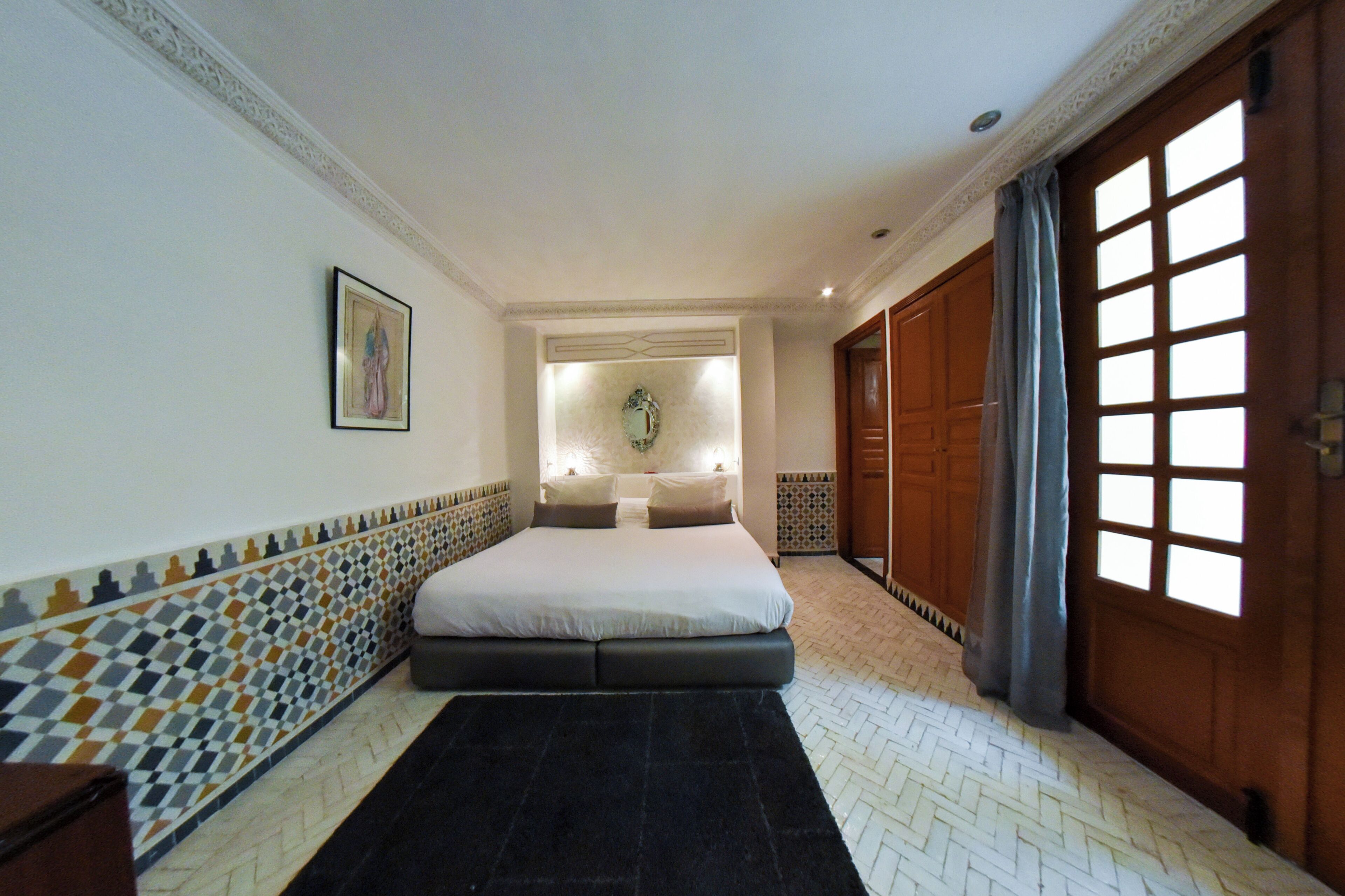 A hotel room with a bed, a nightstand, a mirror, a window with curtains, and a patterned wall.