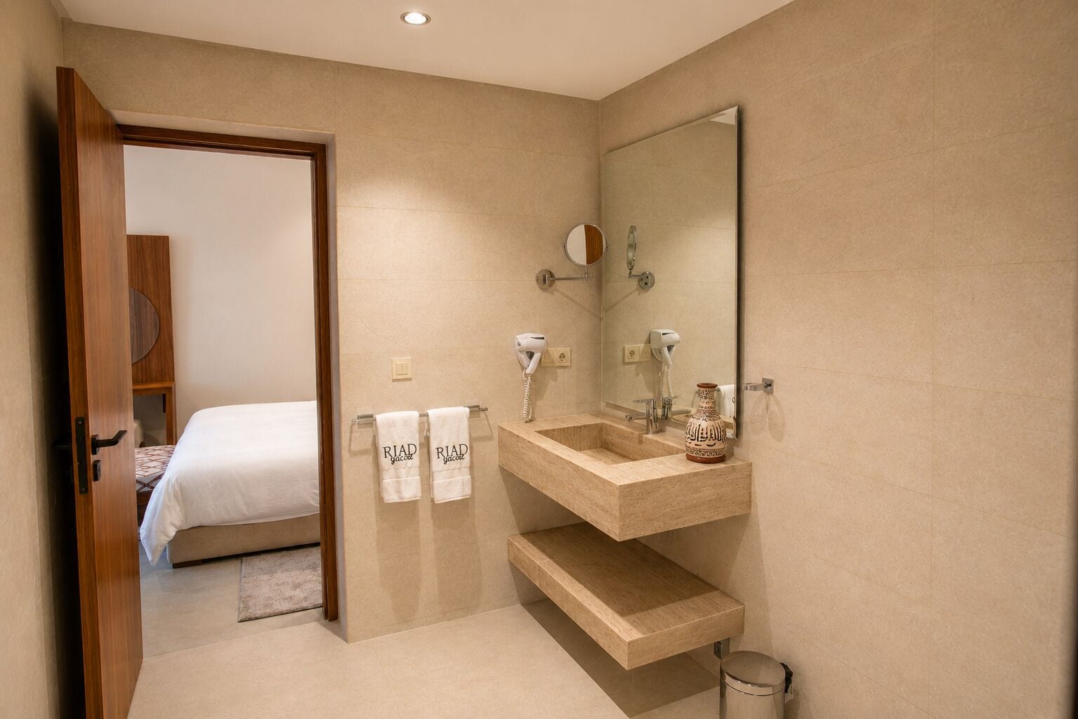 Une salle de bain d’hôtel avec un meuble-lavabo en bois, deux vasques, un miroir et un lit visible à travers une porte ouverte.