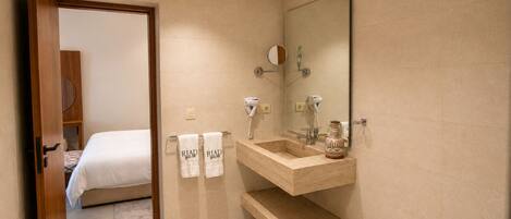 Une salle de bain d’hôtel avec un meuble-lavabo en bois, deux vasques, un miroir et un lit visible à travers une porte ouverte.