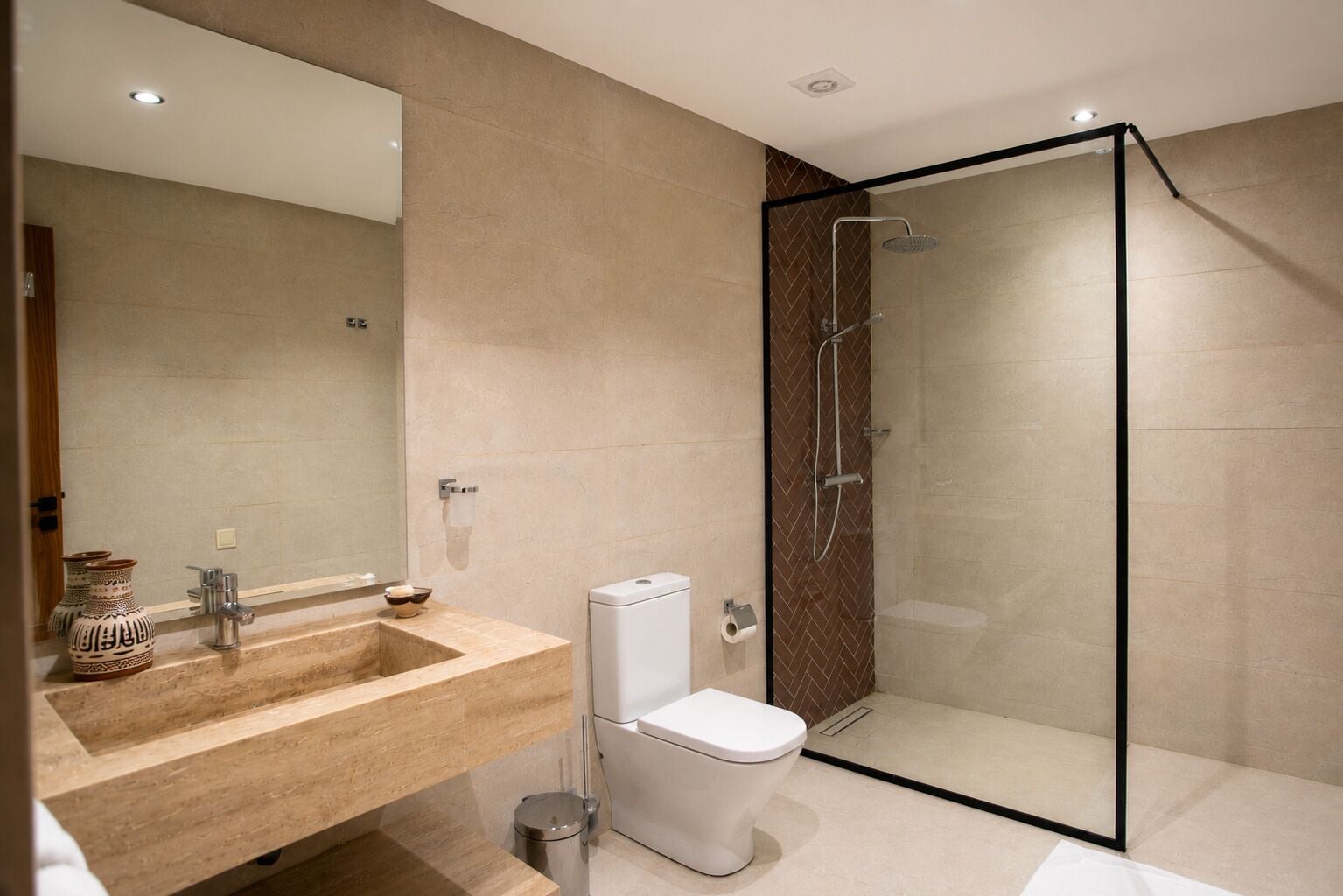 A modern bathroom with a glass-enclosed shower, a beige-tiled sink, and a white toilet.