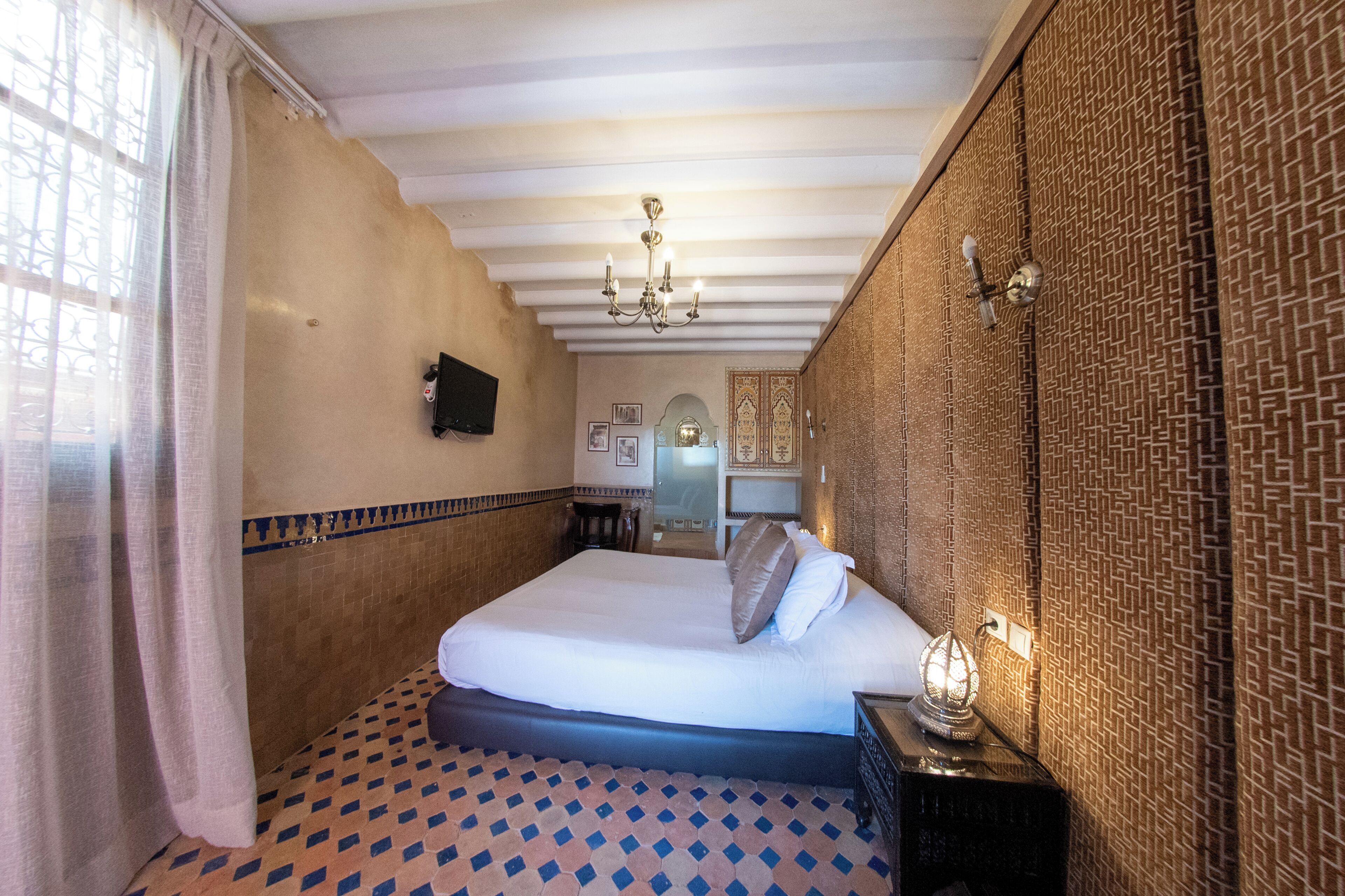 A hotel room with a bed, a nightstand, a wall-mounted TV, patterned wallpaper, and a chandelier.