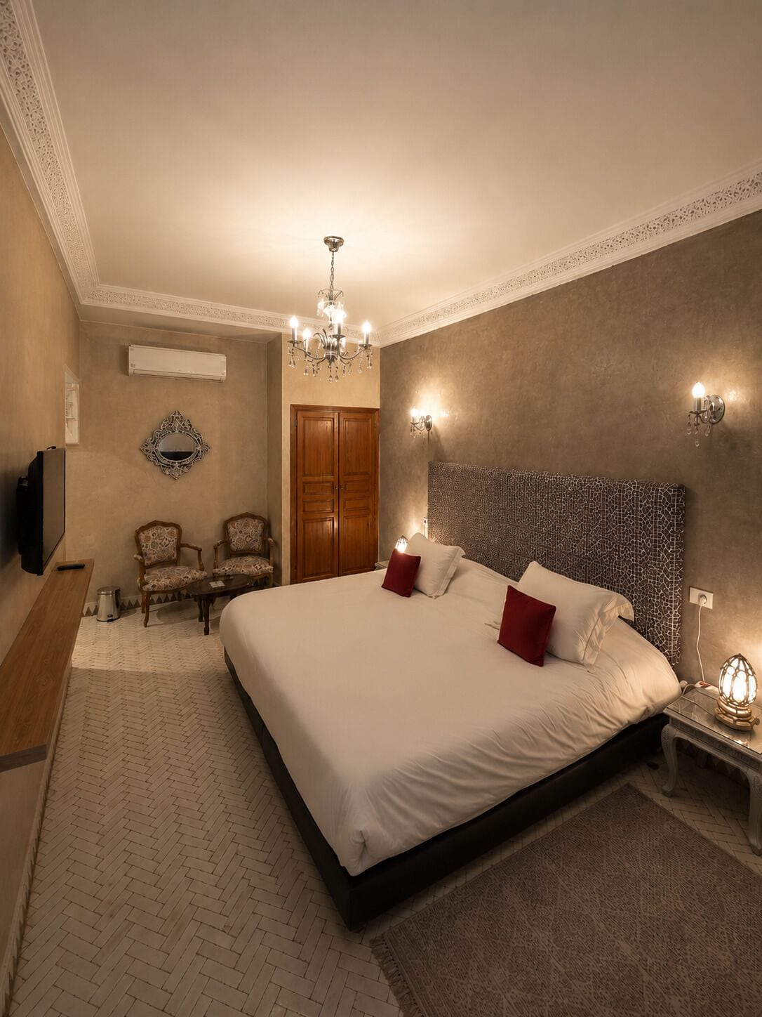 A hotel room with a large bed, a chandelier, a TV, two chairs, a wooden wardrobe, and a patterned wall.