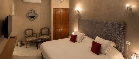 A hotel room with a large bed, a chandelier, a TV, two chairs, a wooden wardrobe, and a patterned wall.