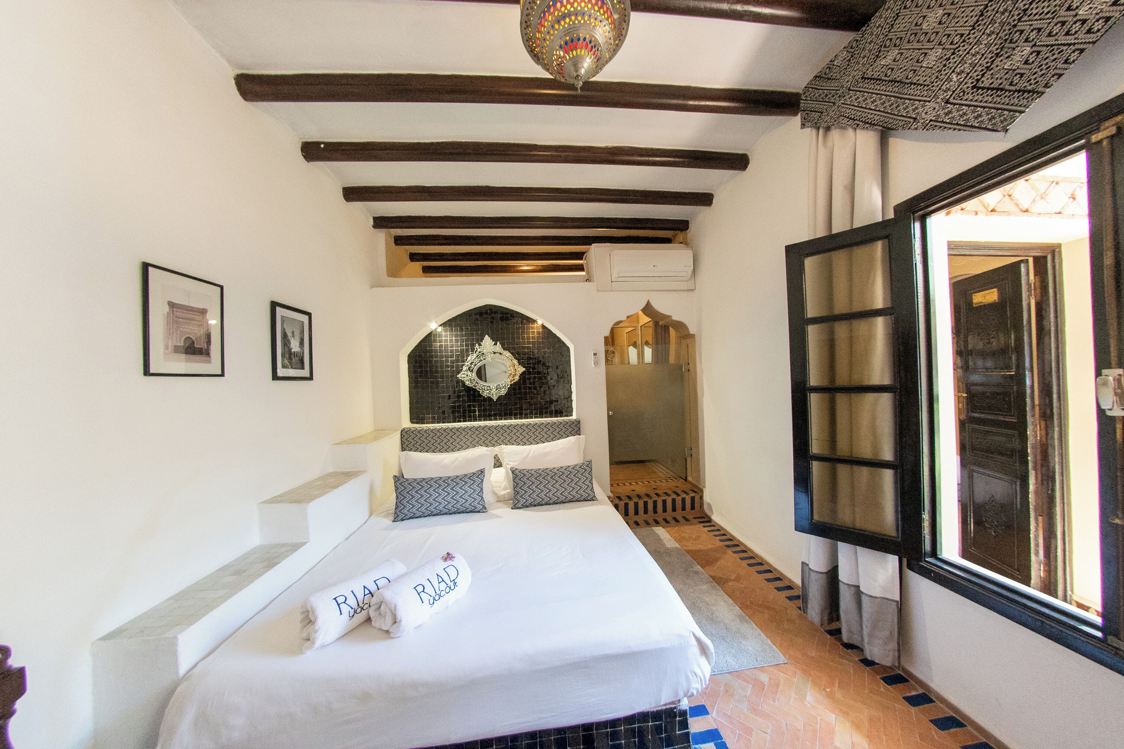 Habitación de hotel con cama, una pequeña escalera, una puerta que da a otra habitación y una ventana con cortinas.