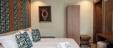 A hotel room with a bed, pillows, a framed artwork, a wooden wardrobe, and a small seating area.