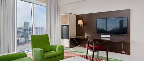 A hotel room with a large window, a green armchair, a white coffee table, a desk with a chair, and a flat-screen TV mounted on the wall.