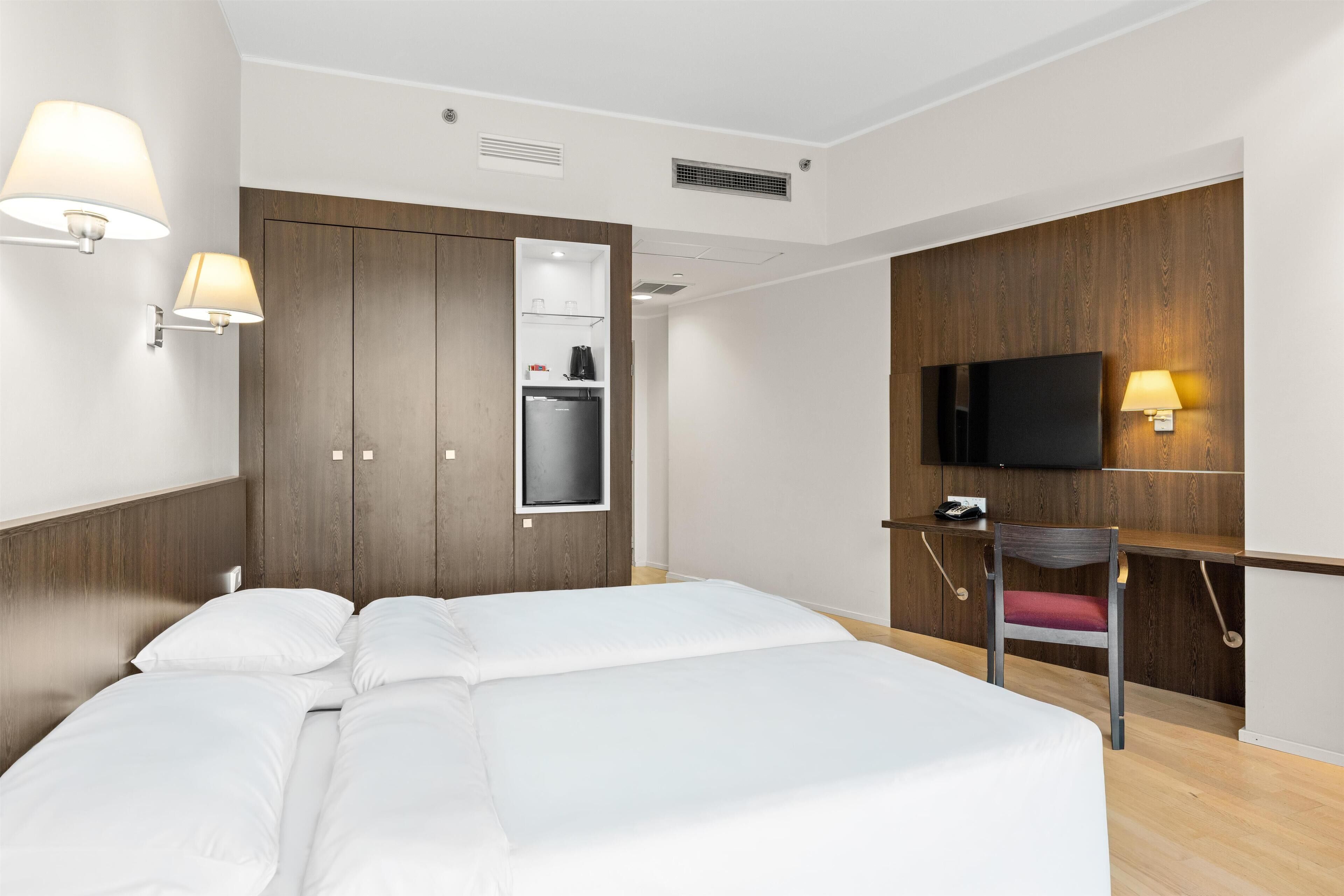 A hotel room with a bed, a wardrobe, a desk with a chair, a television, and wall-mounted lamps.