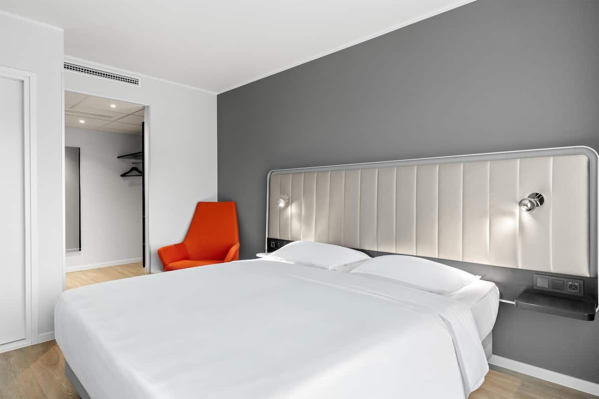 Ein modernes Hotelzimmer mit einem großen Bett, einem leuchtend orangefarbenen Sessel und einer grauen Akzentwand.