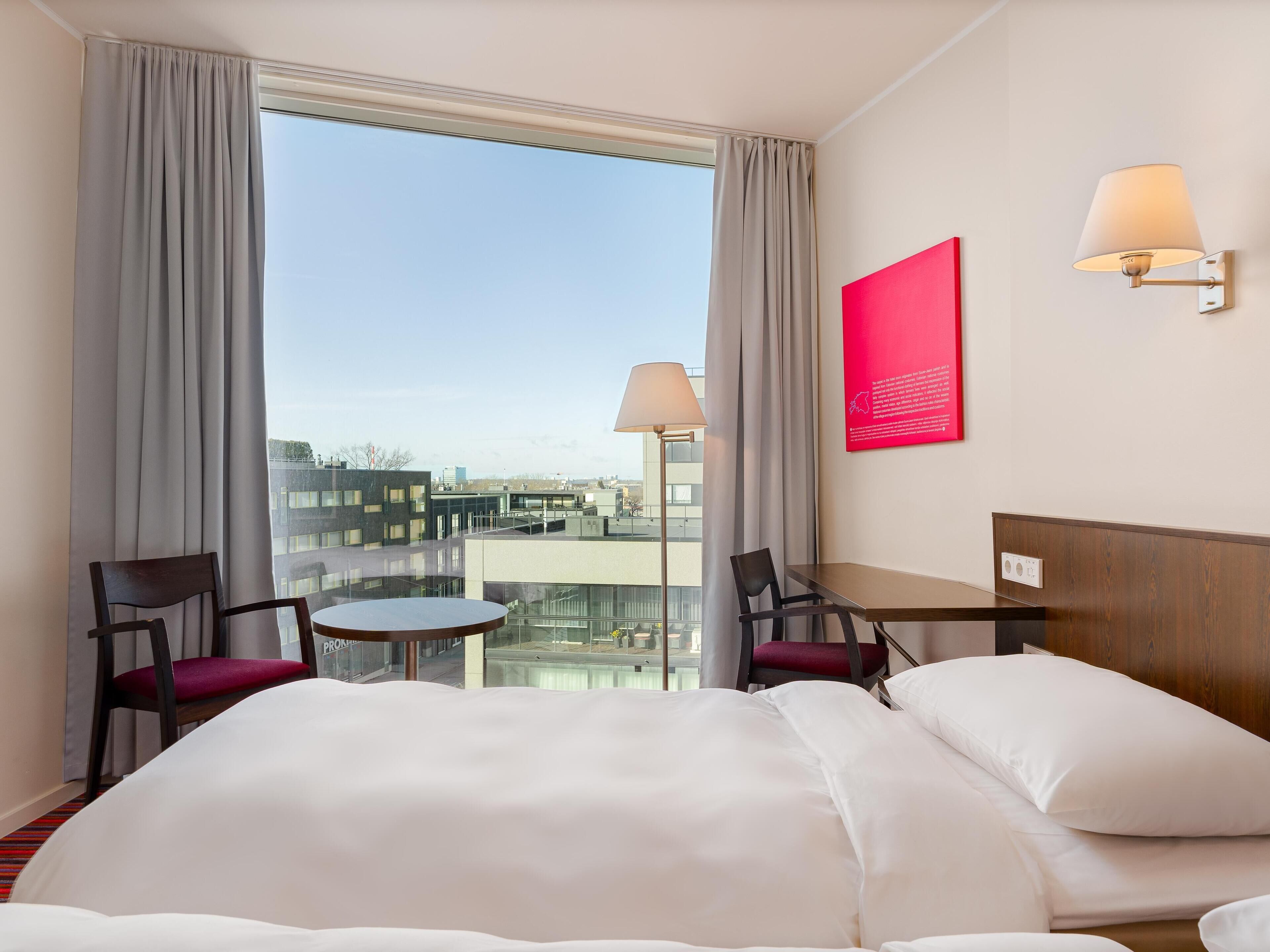 A hotel room with a large bed, two chairs, a small table, a balcony with a view of the city, and a red informational poster on the wall.