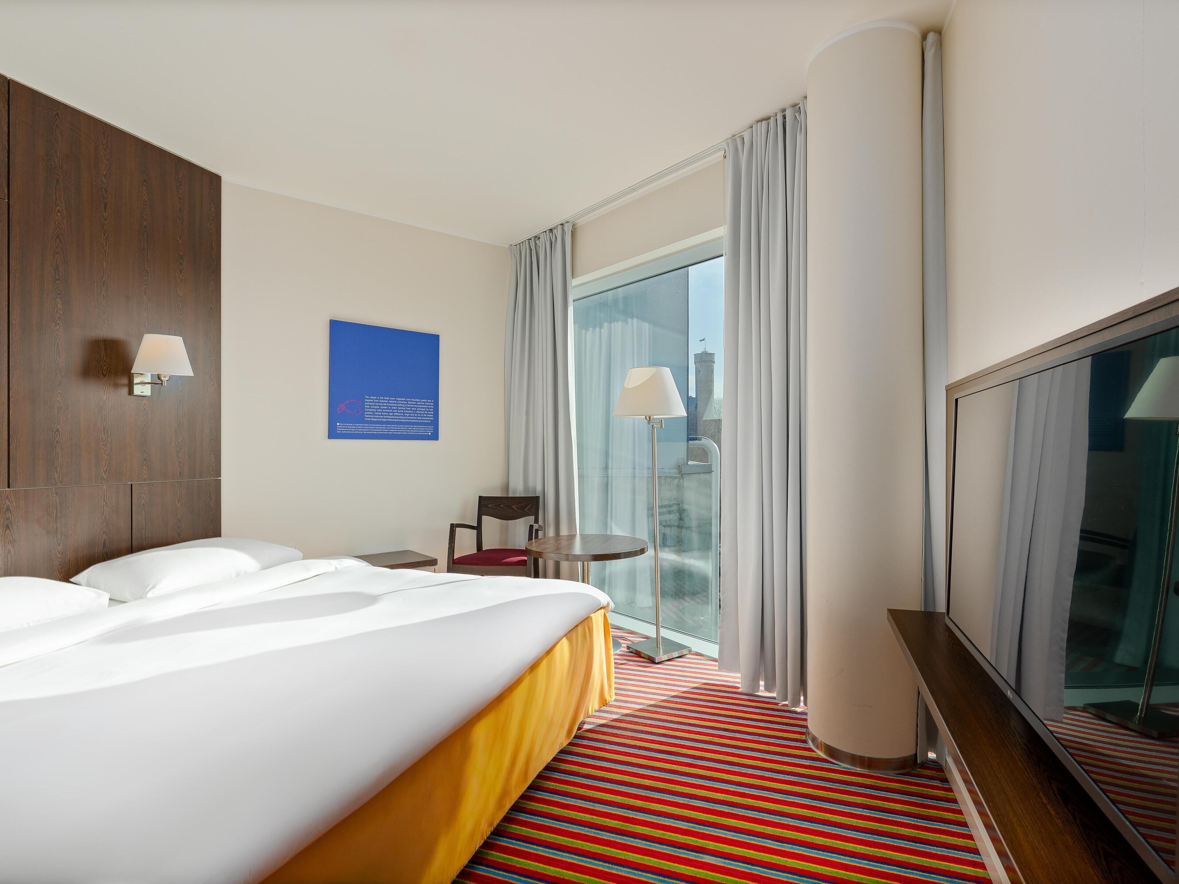 A hotel room with a large bed, a small table, a chair, a TV, and a window with curtains.