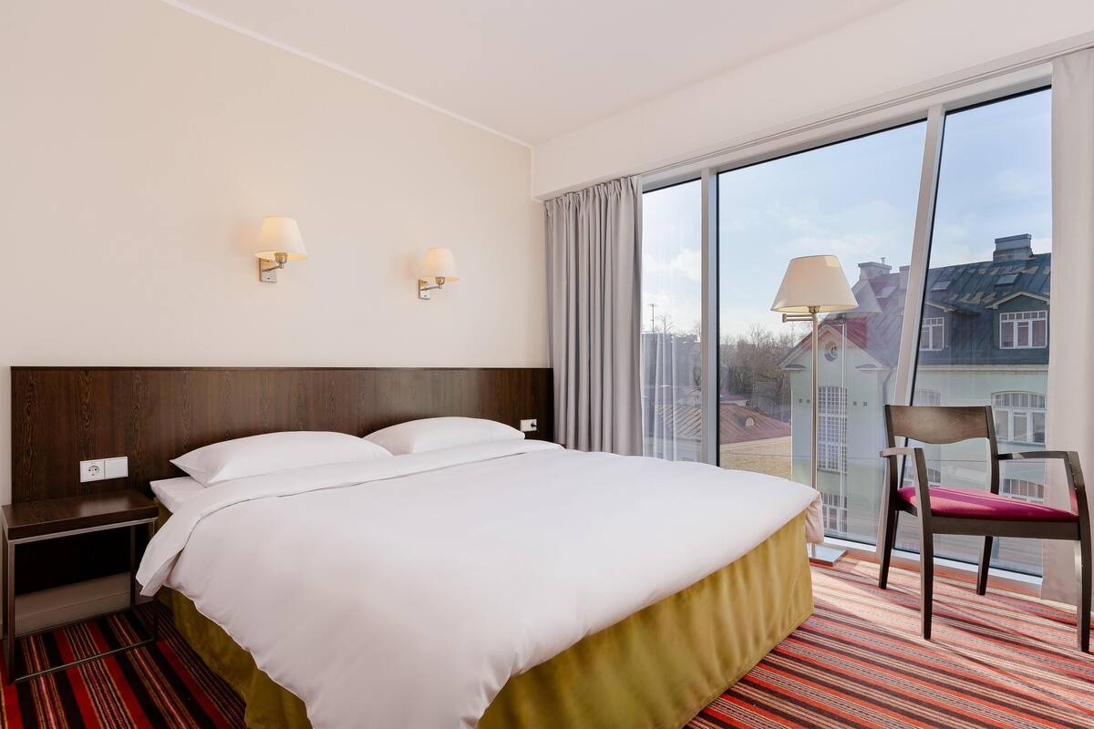 Ein Hotelzimmer mit einem großen Bett, zwei Wandlampen, einem Sessel und einem großen Fenster mit Blick auf die Stadt.