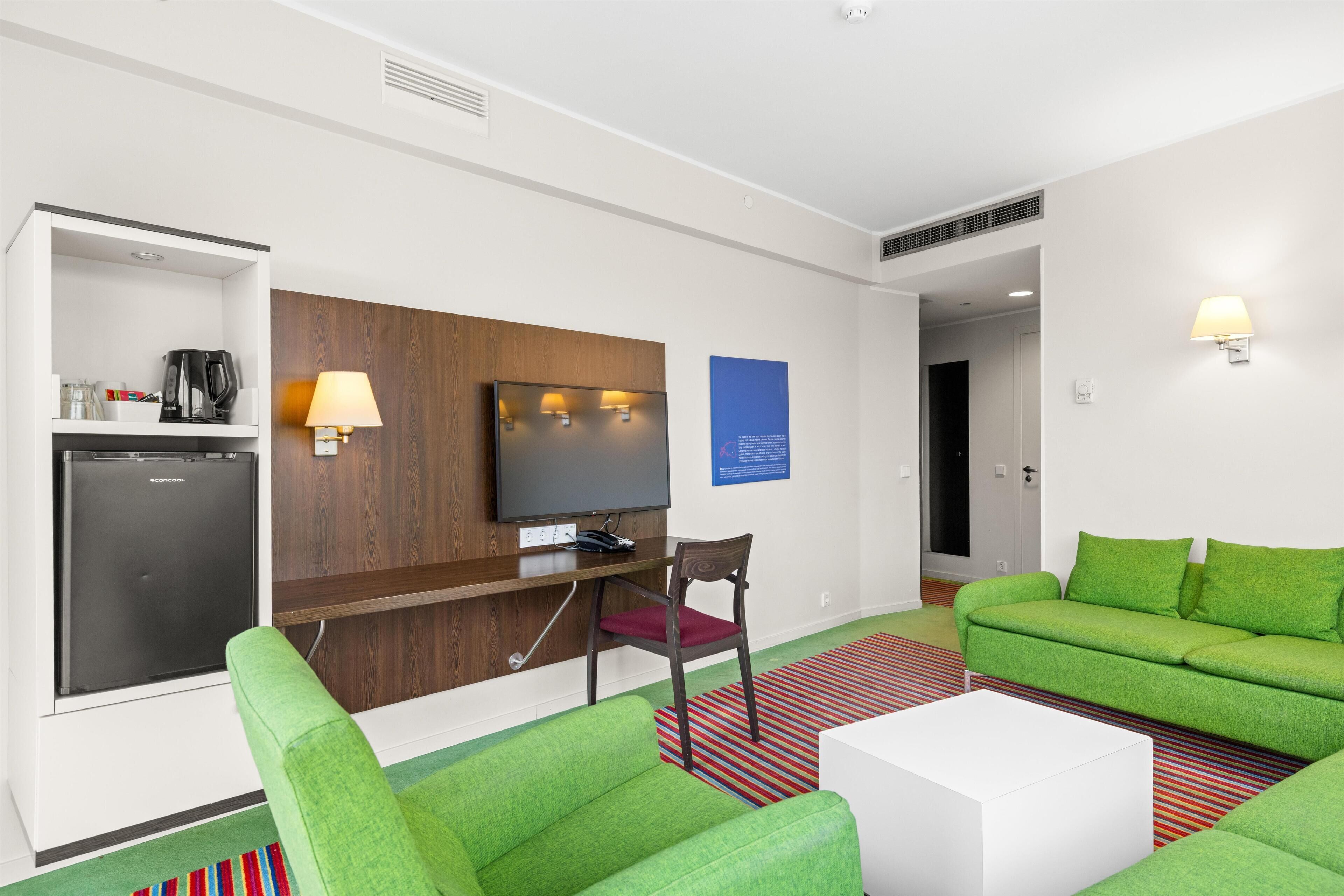 A modern hotel room with a flat-screen TV, a mini-fridge, a green sofa, a white coffee table, and a striped rug.